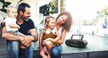Eine Familie mit zwei Kindern sitzt draußen auf einer Terrasse, neben ihnen steht ein Panasonic-Radio.