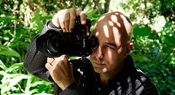 Ein Mann fotografiert mit einer Panasonic-Kamera im Grünen, Sonnenlicht fällt durch Blätter auf sein Gesicht.
